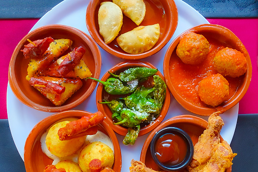 Top view on table of variety of tapas in small clay plates, mushrooms, patatas bravas, chicken in sweet sauce, meat with rice and pumpkin puree served with white sauce, small portions of food