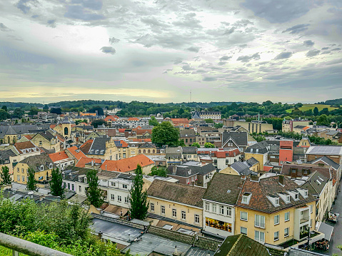 Cityscape of Valkenburg city seen from terrace, buildings with gable roofs in city center, yellow walls, leafy trees on hill in background, cloudy day in South Limburg, Netherlands