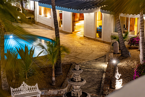 Night view of outdoor rest area in resort with swimming pool, palm trees, quarry fountain, building with yellow lights on, view from balcony, quiet night in Baja California Sur, Mexico