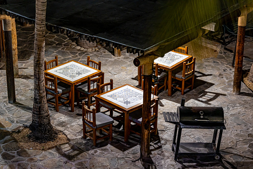 Aerial view of square tables and wooden chairs on terrace of small restaurant, illuminated by light of lamps, stone floor, metal grill, quiet night in Baja California Sur, Mexico