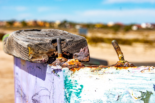 Closeup of two rusty screws and a piece of wood on edge of boat hull against blurred background, peeling purple, green and white paint, sunny day on Mexican beach
