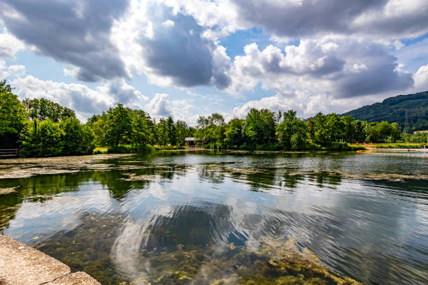 Reflections on water surface in panoramic landscape of Lake Echternach, algae in calm water, hills with abundant leafy trees in background, sunny spring day with abundant clouds in Luxembourg