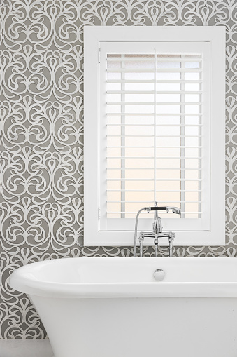 A freestanding soaking tub detail with a grey and white tile pattern wall, polished chrome faucet, and white shutters over the window.