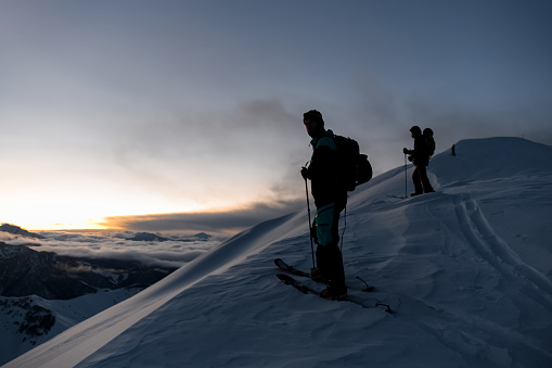Silhouettes of three skiers climbing a mountain slope against the background of lower mountains that are shrouded in fog, all placed at a distance from each other