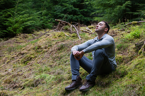 A medium full-length front view of a mid adult male as he takes a moment to reflect and take in the beautiful surroundings whilst on a hike in Northumberland in the North East of England. He is sitting with his eyes closed looking upwards.