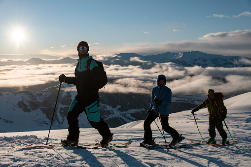 Three male skiers climb a snowy slope against the background of fog-covered peaks and a bright blue sky
