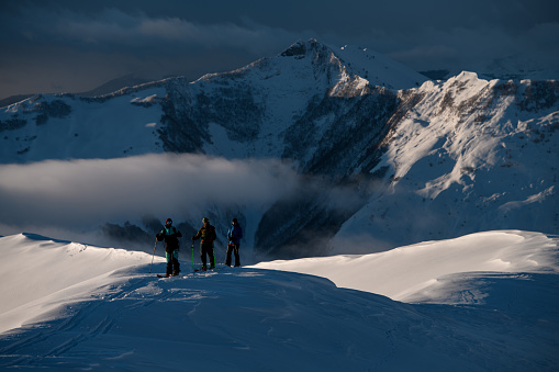 Expedition of three male skiers stand on a snowy slope in full gear against the background of a mountain massif, part of which is covered by a small band of fog