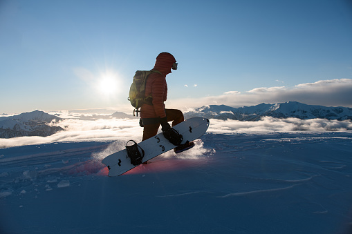 Snowboarder in a red jacket is walking on deep snow, holding his snowboard in his hands against the background of a mountain landscape and a sunny sky