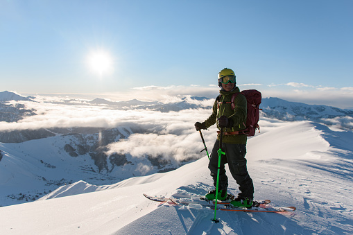 Happy freerider standing on skis on top of highest mountain and admiring other mountain peaks drowning in misty clouds against sunny sky background