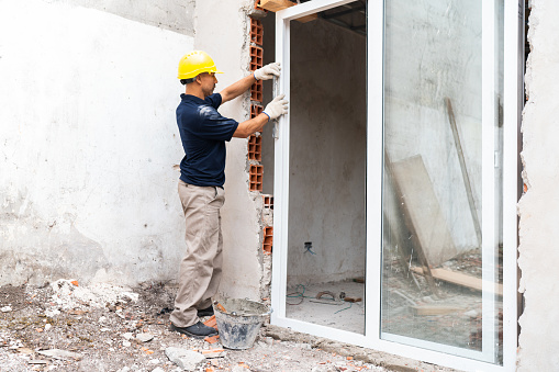 A construction worker carefully installs a new window in a partially built wall at a construction site.