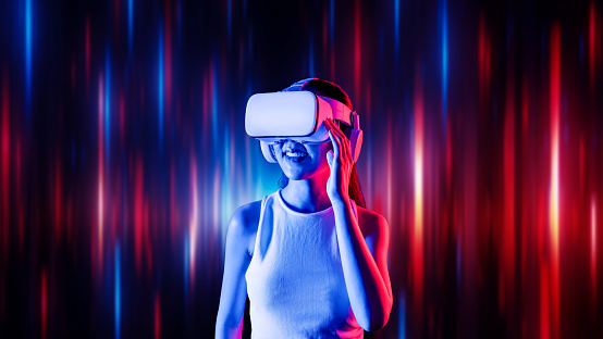 Smart female standing surrounded by neon light wearing VR headset connecting metaverse, future cyberspace community technology. Elegant woman looking faraway and smiling satisfactorily. Hallucination.