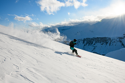 Snowboarder in a black and blue ski suit with backpacks on his shoulders confidently overcomes a route on a smooth snowy slope on his snowboard