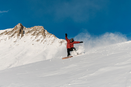 Snowboarder in a red jacket is going down a slope with his hands up for balance against the background of a brown mountain peak