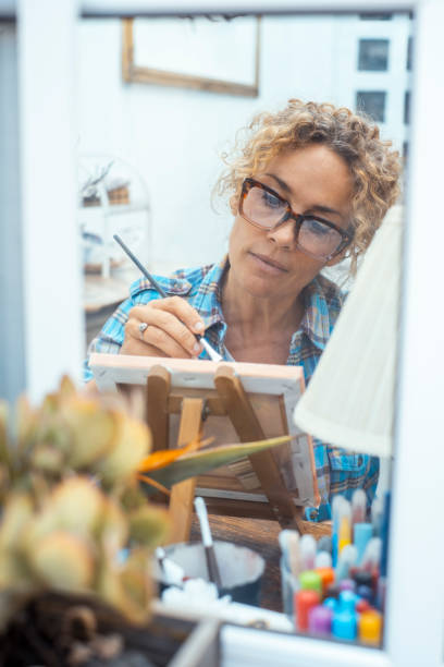 istock Concept of art, creativity, hobbies, work and creative occupation. Front view of blond, curly woman sitting indoors at home in front of a window in front of easel, painting with brush and colors 2086729379
