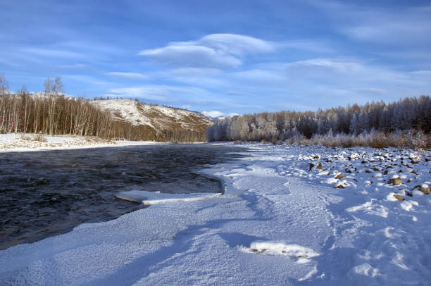 A frozen river surrounded by snow-covered mountains under a cloudy sky. The natural landscape features a sloping terrain and a freezing cold atmosphere. Russia, Chuisky ridge, Katun river.