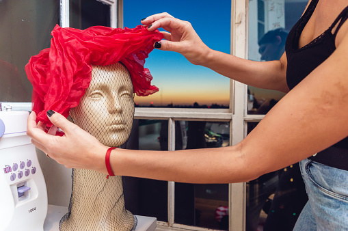 mannequin head on a table by the window wearing a beautiful vintage red female hat, designer arranging the details of the vintage hat placed on the mannequin