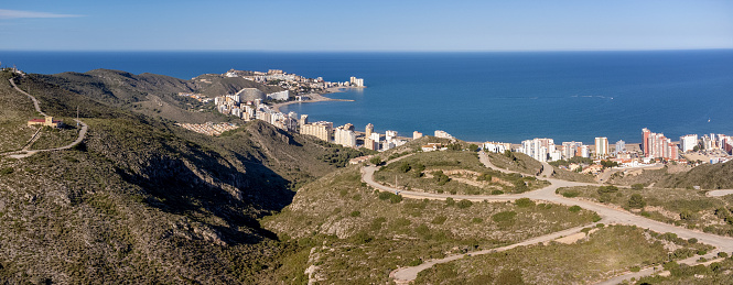 A scenic landscape photograph capturing the coastline of Cullera with the Cullera mountain range in the foreground. In background the summer apartment buildings along the beachfront. Travel concept in Spain