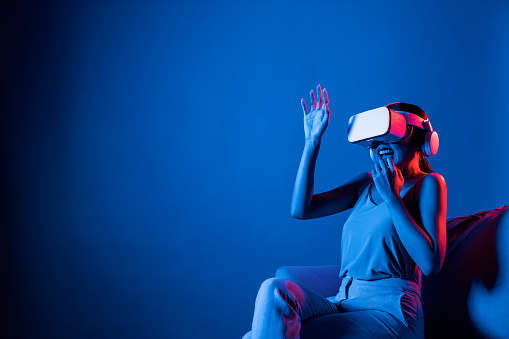 Smart female sitting on sofa surrounded by neon light wear VR headset connect metaverse, futuristic cyberspace community technology. Elegant woman excited and emotionally watch movie. Hallucination.