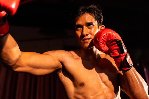 Boxer punch his fist in front of camera in ready to fight stance posing at gym with boxing equipment in background. Focused determination eyes and prepare for challenge. Impetus
