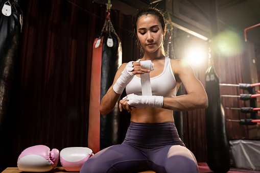 Determined Asian female boxer with muscularity physical readiness body wraps her hand and dons or wearing boxing glove, preparing for intense boxing training in the ring at gym. Impetus
