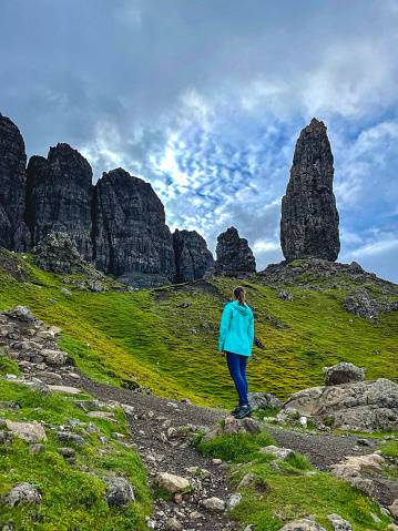 Young lady stopped on a hiking trail to look up at the majestic Old Man of Storr. An imposing rock pinnacle stole attention of a tourist climbing towards a famous natural landmark on the Isle of Skye.