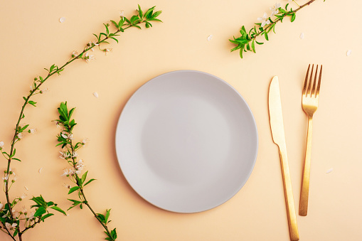 Gray plate and cutlery on light peach background with cherry blossom branches. Top view, flat lay, mockup.