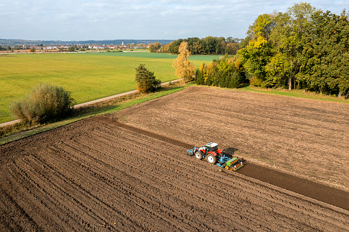 Aerial view of a red, agricultural tractor cultivating and sowing plowed field on a sunny day in a rural landscape.