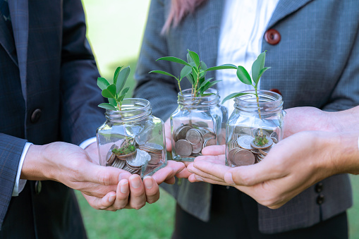 Concept of sustainable money growth investment with glass jar filled with money savings coins with businesspeople as eco-friendly financial investment nurtured with nature and healthy retirement. Gyre