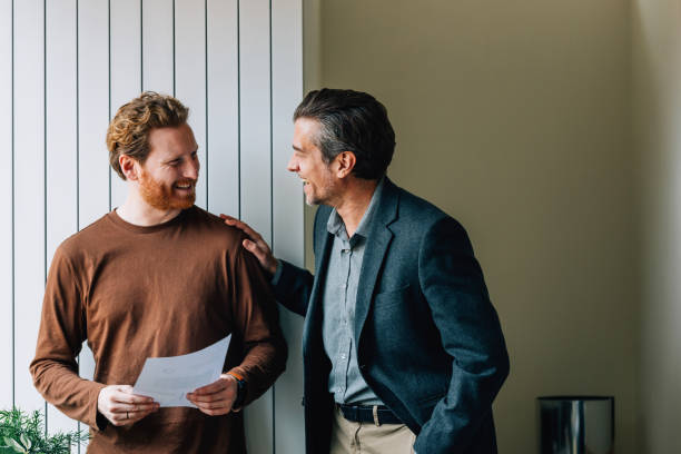 uśmiechnięty młody mężczyzna i starszy mężczyzna z dokumentem omawiający w biurze - codzienne-ubranie zdjęcia i obrazy z banku zdjęć