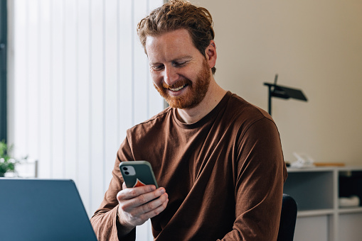 Joyful businessman browsing his smartphone at a desk with a laptop in a bright office setting.