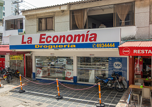 Cartagena, Colombia - July 25, 2023: La Economia Drogueria, pharmacy and beayty products retail business along Calle 29. \nFacade with colorful sign. Motorbikes and Coca-Cola sign at restaurant Lancem