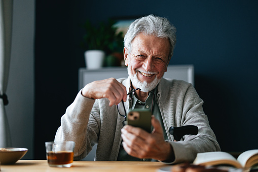 A smiling old man holding eyeglasses and his smartphone while looking at camera and sitting at the table with a book, cup of tea and biscuits.