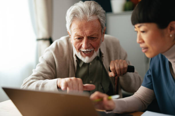 Worried Senior Man Using A Laptop Computer And Paying Bills With An Anonymous Nurse
