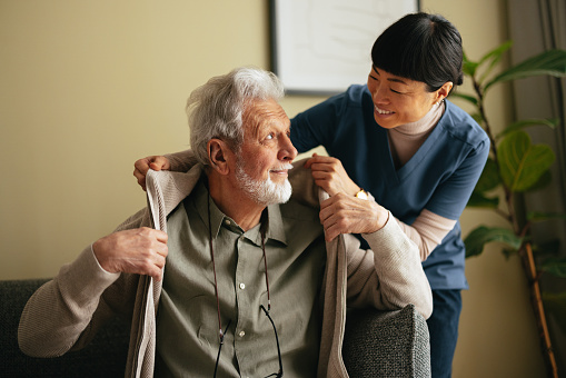 Close up shot of a smiling elderly man sitting on the sofa and a beautiful Asian female carer. She is smiling while helping him put on or take off his jacket.