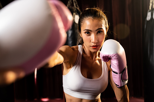 Asian female boxer punch fist in front of camera in ready to fight stance posing at gym with boxing equipment in background. Focused determination eyes and prepare for challenge. Impetus