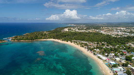 A stunning aerial coastal landscape, featuring a clear blue sea adjacent to a curved sandy beach. The beach is bordered by Cerro Gordo, a coastal town on the north shores of Puerto Rico near Dorado Beach.