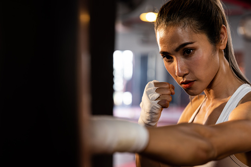 Asian female Muya boxer training, wrapped hand punching at kicking bag at the gym. Healthy sport and fitness lifestyle, Strength and stamina training for boxing match. Impetus