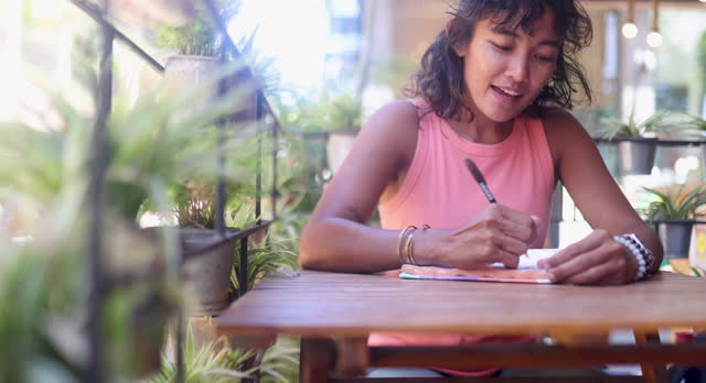 An Asian woman is writing something in a notebook while waiting for a cup of coffee to be brought to her in a coffee shop