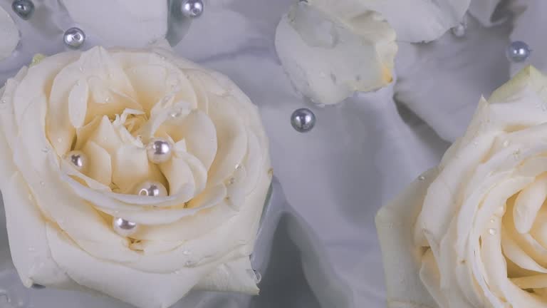 Flowers and petals of white roses with pearls float on the surface of the water on a gray silk background.