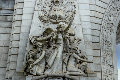 “Spirit of Commerce” is a high relief allegorical sculpture on the north side of the triumphal arch on the Manhattan side of the Manhattan Bridge. The figures are all larger than life. Industry is represented by a dynamically gesturing winged female figure. A male figure in a helmet holding a heavy tool crouches on her on her right, and a female holding a large gear is on her left. \n\nThe sculptor was  Carl Augustus Heber (1871-1956,) and the work was completed in 1914. \n\nHeber’s model for “Industry” was Audrey Munson.  Munson posed for more than a dozen important statues, sculptures, and friezes, most of which still stand to this day across Manhattan and Brooklyn. She is now often called “America’s First Supermodel.”