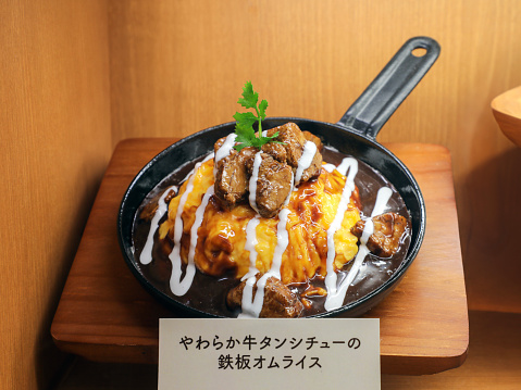 A delectable skillet omelette topped with hearty beef stew, garnished with a drizzle of creamy sauce and a sprig of fresh parsley, displayed as a food model, showcasing a fusion of Western and Japanese cuisine.