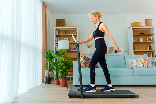 young Woman Walks on a Treadmill and Uses a Tablet at Home