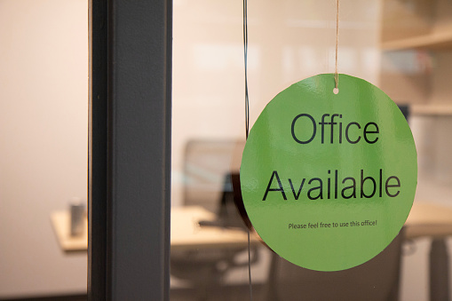 Office available sign shown from the outside of office door at a modern workplace with open seating.