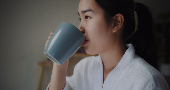 Young asian woman in bathrobe enjoying a healthy drink in wellness resort, She holding a mug in hand Raise and inhale the aroma before drinking, and smiled with satisfaction