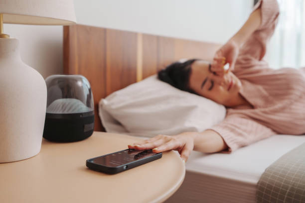 Woman in distress resting in bed, soft light illuminating the room Exhausted young Asian woman in a pink sweater lying in bed, reaching towards a phone on the bedside table alarm clock stock pictures, royalty-free photos & images