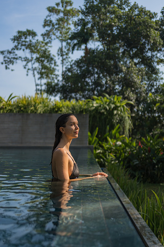 Young woman relaxes on edge of outdoor swimming pool in tropical jungle