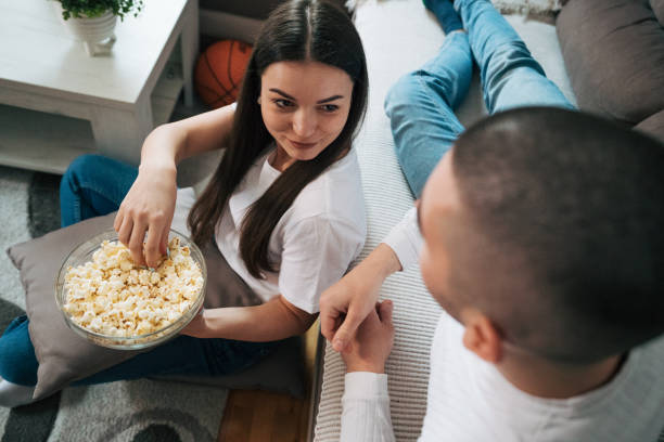 istock A young couple is sitting at home and talking while snacking on popcorn. 2088141265