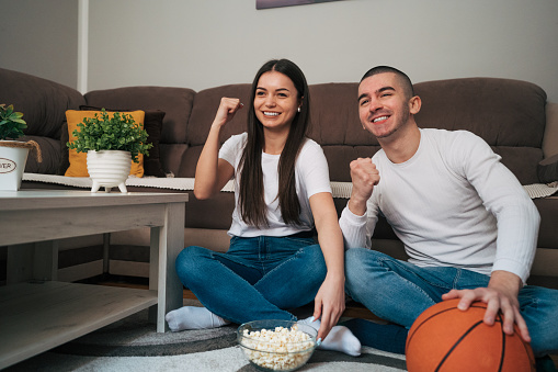A young happy couple is sitting on the floor in their comfortable home and watching a basketball game.