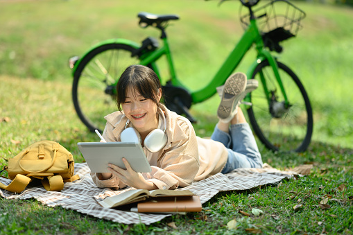 Smiling young woman lying on blanket in the public park and using digital tablet.