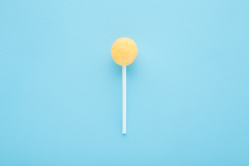 Yellow lollipop on stick on light blue table background. Pastel color. Sweet candy. Closeup. Top down view.